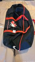 Photo of a black Nike hockey bag with red handles and red trim, showing the Team Canada logo and Nike swoosh logo, laid flat on a beige surface.