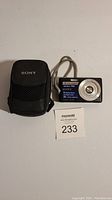 Sony digital camera and black carrying case, laid on white background with MaxSold lot number card near them