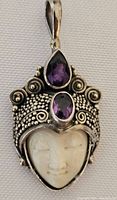 Front view of pendant with carved face and intricate silver detailing, two purple stones on top.