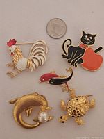 Five animal brooch pins displayed together with a coin for scale including a rooster, cat with apple, dolphin with pearl, two fish, and a turtle.