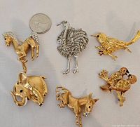 Five animal brooch pins arranged with a quarter coin for scale: horse, peacock, bird with colorful eyes, elephant, and two small birds on a branch with rhinestones.