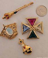 All four goldtone royalty-themed brooches with glass stones, including scepter, crown, Maltese cross, and orb and cross pieces, shown against a plain background with a coin for scale.
