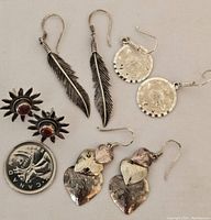 Four pairs of earrings displayed with a Canadian quarter for size reference. Includes feather drop earrings, hammered disc drop earrings, heart drop earrings, and sun-shaped stud earrings with red stones.