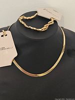 Gold-tone Gloria Vanderbilt necklace and bracelet displayed on a black stand, showing the necklace's smooth flat chain and the bracelet's twisted link design with original tags.