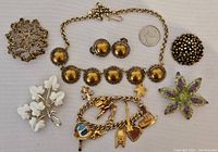 Full view of all seven jewelry pieces in the lot arranged on a beige cloth background with a Canadian quarter for scale: gold-tone necklace with round golden cabochon stones, charm bracelet with multiple charms, and various brooches.