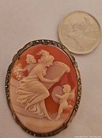 Front view of an oval shell cameo brooch pendant showing a classical scene with women and a cherub, alongside a coin for size reference.