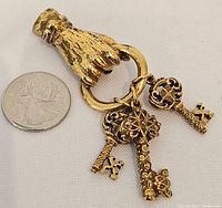 Front view of brooch showing textured gold-tone hand holding ring with three ornate keys hanging.