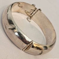 Angled view of vintage sterling silver bangle bracelet showing polished surface, hinge clasp, and attached safety chain.