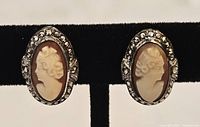Front view of two oval cameo earrings featuring shell cameos with female profiles and rhinestone borders on a black stand.