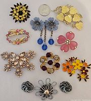 All ten jewelry pieces laid out on white cloth with a quarter for size reference. Shows full collection including floral brooches and clip-on earrings in varied colors.