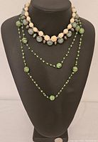 Image shows three vintage necklaces displayed on a black bust, two made of glass beads in shades of green and one cream necklace with carved resin rose beads and speckled beads.