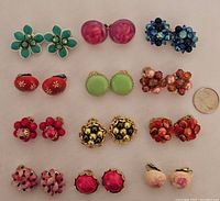 Full set of twelve pairs of vintage Hong Kong clip-on earrings on white fabric background, showing various colors and designs including flowers and beads.