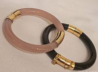 Pink and black quartz bangles positioned side by side, showing their circular shape and gold-tone metal ends with safety chains.