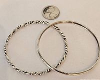 Two silver bangles placed next to a quarter showing scale. One bangle is twisted rope design; the other is a fine beaded texture design.