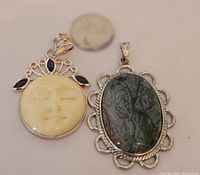 Both pendants displayed side by side with a quarter for size comparison. The moon face pendant is cream-colored with purple stones, the stone set pendant is dark green with silver scalloped edges.