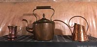 Front view of all three copper items: kettle, pitcher, and watering can on a surface.