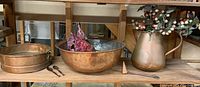 Wide view of shelf showing large copper sifter, large hammered copper bowl with artificial flowers, tall copper water pitcher with artificial berries, small copper candle snuff, and two wooden spoons arranged on wooden shelf.