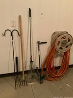 Full view of gardening tools and hose reel against white wall, showing black double hook shepherd's hook, pitchfork, rake, weed puller, and coiled orange hose on gray reel.