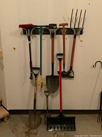 Full view of the seven long handled tools hanging on a rack on a wall including two shovels, cultivator, garden fork, snow brush, edger, and hoe.