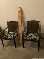 Pair of dark brown plastic wicker patio chairs with metal frames and tropical leaf cushions beside unopened umbrella box.