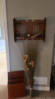 Photo showing the vintage Art Deco style wood wall shelf mounted on the wall with decorative items including the wedgewood-style clock and wall hanging shoe placed on it. Below are the wood magazine holder and a tall bundle of faux florals leaning against the wall.