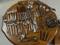 Full view of flatware spread on round wooden table showing assortment of forks, knives, spoons, serving utensils, and salt and pepper shakers.