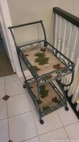 Full side angle of metal framed tea cart with two resin trays decorated with leaf patterns, sitting on white tile floor next to stair railing.