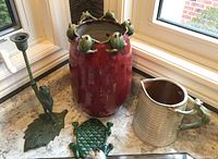 Overview of the pottery vase with frogs around the rim, pottery jug with frog handle, metal frog trivet, and frog candle holder grouped together on a surface.