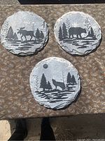 Three round concrete composite plaques displayed showing moose, bear, and wolf silhouettes in landscape settings.