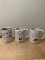 Front view of the three Rae Dunn Easter mugs showing text and rabbit illustrations, placed side by side on a wooden surface.