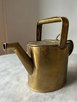 Side angled view of the brass watering can showing the handle, spout and body with patina and minor wear.