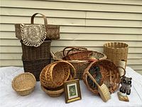 Front view of the lot displaying various sizes and styles of woven baskets, owl figurine, small framed picture, treen brush, and wooden spool holder with thread.
