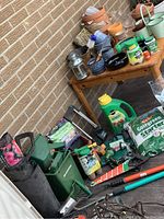 Wide view showing large Rubbermaid tub, assorted garden hand tools, flowerpots, spray nozzles and fertilizers on wooden bench and ground