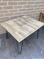 Front side view of square hardwood tabletop with black hairpin legs on patio floor beside brick wall.