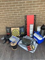 Overall lot showing mechanic creeper, dart board with darts, multiple tie down straps with ratchets, coolers, thermal beverage containers, ammo box, spool of cord and silver tarp.