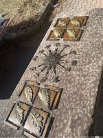 Three metal wall plaques displayed flat on fabric surface outdoors: central circular zodiac astrology wheel with sun and signs, two square foliate leaf plaques each with four raised leaf designs.