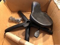 View inside box showing black 5-legged plastic base with pedals and black contoured seat, packaged with some wrapped components for assembly.