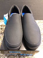 Front view showing both black slip-on shoes on box, with tags attached.