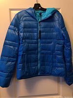 Front view of blue women's puffer jacket with full zipper and hood, hanging on a hanger against a door.