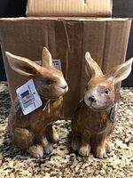 Two ceramic bunny figurines shown from front with property tags, against a kitchen counter and cardboard box background.