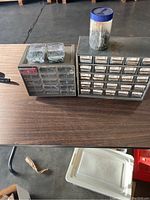 View of two metal hardware cabinets on a table, one with 15 drawers and the other with 25 drawers, both filled with assorted screws and nails. Additional containers filled with screws placed on top of cabinets.