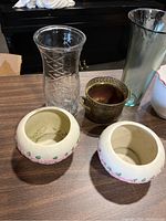 Photo shows all six vases together on a wooden table: two small ceramic vases with floral detailing, a brass vase with perforated design, and three glass vases including a clear diamond-patterned one and taller tinted ones.