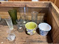 Photo showing all 9 vases arranged inside a wooden crate, displaying variety in glass and ceramic materials and shapes.