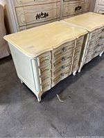 Two cream-colored vintage nightstands or small dressers with curved drawer fronts and decorative metal drawer pulls, showing surface scratches and worn spots.