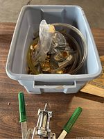 Photo of gray plastic tote containing assorted bottle caps and brewing tubing, with metal bottle capper having green handles placed in front