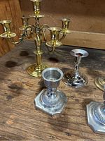 Group of brass and metal candle holders and candelabras placed on a wooden surface, showing variety of designs and bases.