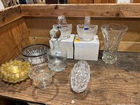 Full view of all crystal serving vessels and vase on wooden surface including footed dish, oval dish, two square handle dishes, decanter, tall vase, small yellow bowl, and clear round dish.
