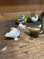 Photo showing five bird figurines on a wooden surface, including a brass swan, white ceramic duck, white ceramic bird, and a colorful blue bird in a nest.
