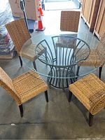 Full view of the round dining table with clear glass top and four woven chairs around it for scale.