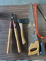 Photo of pruning lopper, small hatchet, hand saw with wooden handle, pipe wrench, and edge of other tools lying on a dark wooden surface.